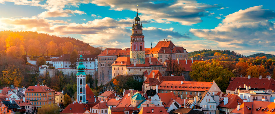 Autumn townscape with red-tiled roofs, a tall church tower, and a castle on a hill beneath a blue sky.